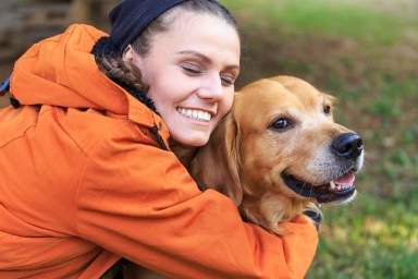 Smiling young woman kneeling and embracing a dog. Wears casual clothes, with eyes closed. Focus on foreground.
