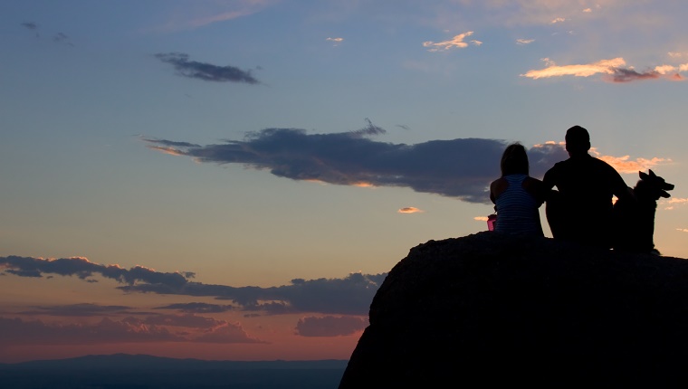 A couple sit with their dog on a rock looking at the sunset over the mountains in the distance.