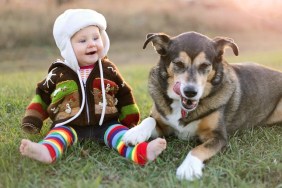 An adorable 8 month old baby girl is bundled up in a sweater and wearing a winter earflap hat looking lovinlgy at her pet German Shepherd dog as they sit and laugh ouside on a cold fall day.