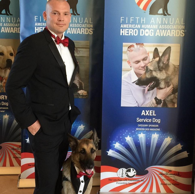 Jason and his German Shepherd service dog, Axel, stand in front of a banner for the American Humane Association's Hero Dog Awards.