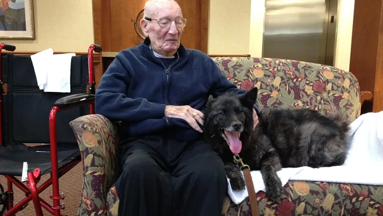 An elderly man sits on a couch with a black dog.