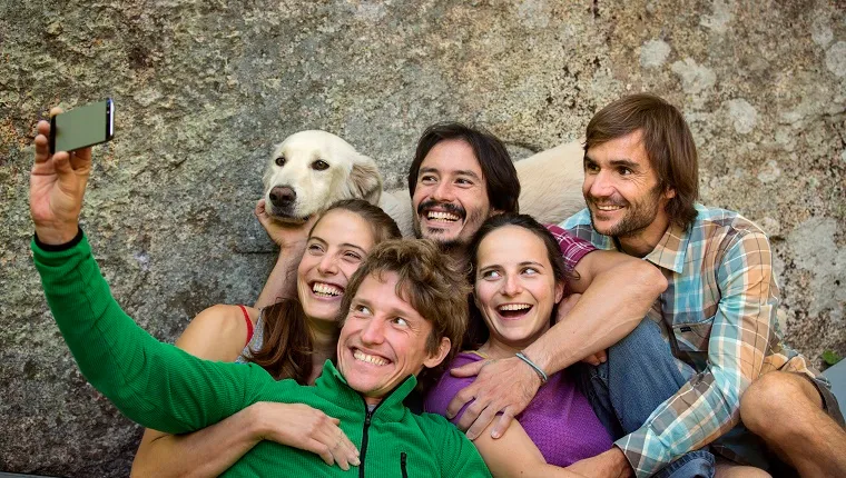 A group of hiking friends take a selfie with a Golden Retriever in front of a rock.
