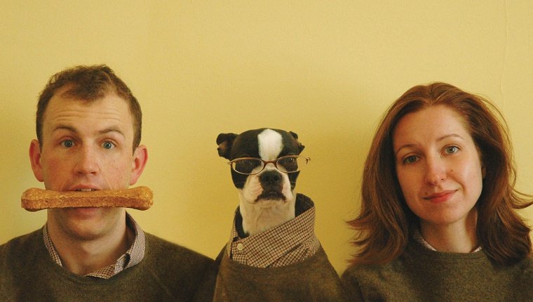 Humorous Christmas card portrait of man, woman, and Boston Terrier pet dog. Dog takes on human attribute of wearing glasses, man in turn has dog bone in mouth. Woman is bemused by entire affair. Family dressed in matching sweaters and shirts.