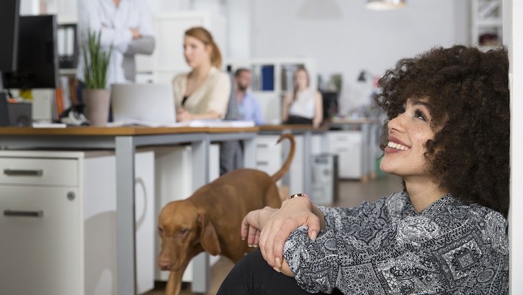 Smiling woman in office with colleagues and dog in background