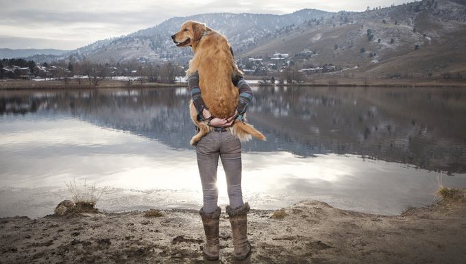 man holds dog near lake and mountains
