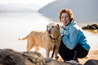 A smiling young woman with red hair and a blue jacket sits next to a golden labrador on the shores of Priest Lake. Priest Lake, Idaho.