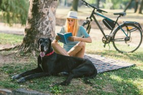 Mid adult woman enjoying the summer day with her dog, great dane, in the park. They are sitting on blanket and woman is reading book.