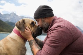 Affectionate bearded man with tattoos kissing dog at remote lakeside