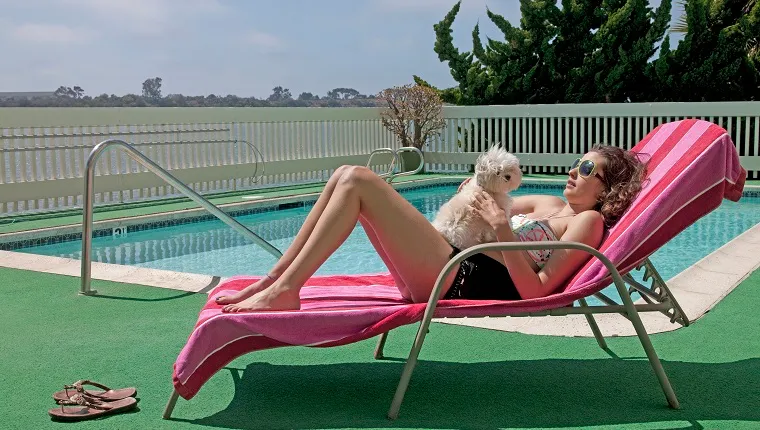 Woman laying down on lounge chair by swimming pool and holding dog on her stomach