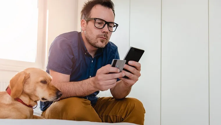 Man with glasses in blue shirt sitting on the bed and using smart phone to pay with credit card.