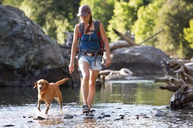 A hiker and her dog cross the shallow part of a river in the western United States. They are on a day hike and the woman is carrying a small backpack.