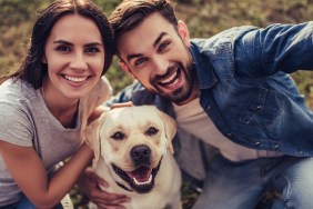 Beautiful romantic couple is having fun with their dog labrador retriever outdoors. Sitting on a green grass and making selfie.