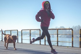 Young fit woman accompanied with her dog, running and listening to music on the riverbank in the city. The woman wears black running tights, yellow hat and red jacket. She has a smart phone mounted on her arm, with earphones connected to it. Her body is in motion, slightly above the ground. The copy space has been left. Shallow DOF.