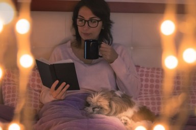 Young Woman Holding Mug Reading Book While Sitting On Bed At Home