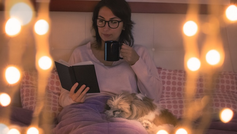 Young Woman Holding Mug Reading Book While Sitting On Bed At Home