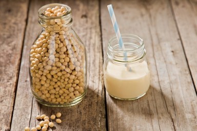A close up shot of an old milk bottle filled with dry soy beans with a few beans on the foreground and a small glass jar container with soy milk and a straw. Shot on an old grungy wooden table.