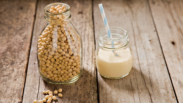 A close up shot of an old milk bottle filled with dry soy beans with a few beans on the foreground and a small glass jar container with soy milk and a straw. Shot on an old grungy wooden table.