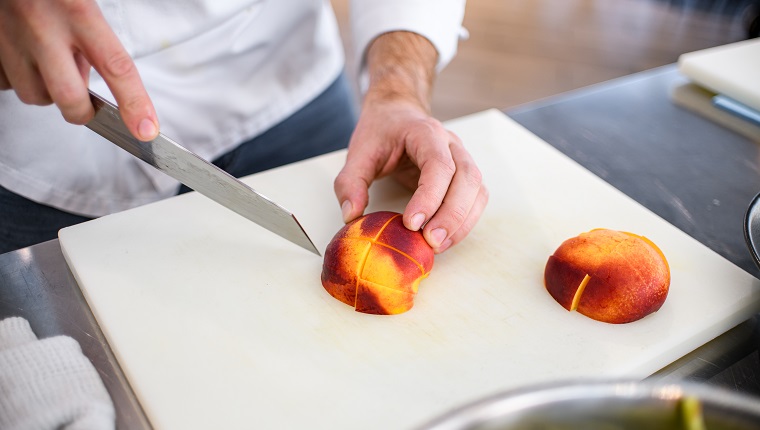 view of ripe peach fruit sliced on a white cutting board and male chef hand with sharp knife nearby