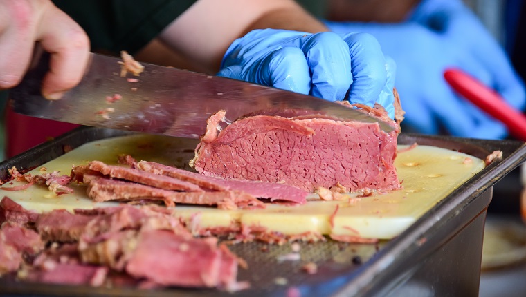Close up of a hand wearing protective glove holding and slicing pastrami on a cutting board at a food market