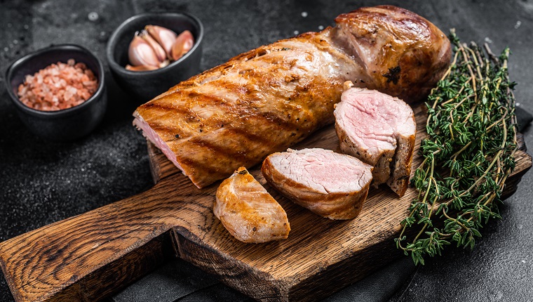 BBQ roasted pork tenderloin fillet meat on wooden board with herbs. Black background. Top view.