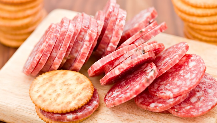 A high angle close up shot of a wooden cutting board with sliced salami and a slice of salami between two crackers. Two stacks of crackers sit on the foreground