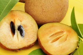 Sapodilla, Manikara Zapota, is a popular fruit in Mexico, South America, the Caribbean, and Middle Eastern countries like India and Pakistan. Also known as sapota, zapota, and chikoo. This image shows a sliced fuit in foreground with seeds in one side. A whole, uncut fruit is in the background, The fruit is on a yellow platter and surrounded by several sapodilla tree leaves. Fruit is very sweet when ripe and has a malty, caramel flavor.