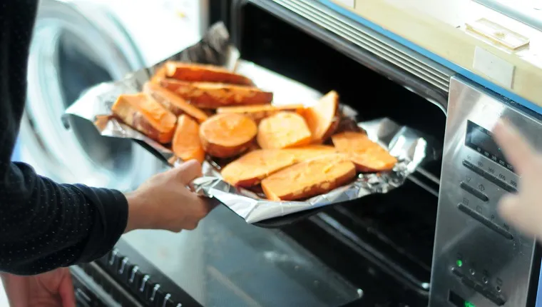 A woman putting sliced sweet potatoes on a baking tray lined with tin foil into an open oven