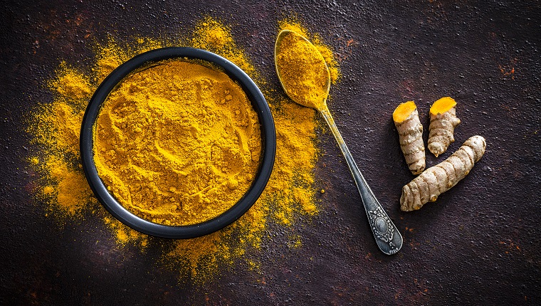 Spices: Top view of a black bowl filled with turmeric powder shot on abstract brown rustic table. A metal spoon with turmeric powder is beside the bowl and turmeric powder is scattered on the table. Fresh organic turmeric roots are beside the spoon. Predominant colors are brown and yellow. Low key DSRL studio photo taken with Canon EOS 5D Mk II and Canon EF 100mm f/2.8L Macro IS USM.