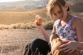 A girl smiles and pets her dog's head with one hand as she holds a bagel in the other hand.