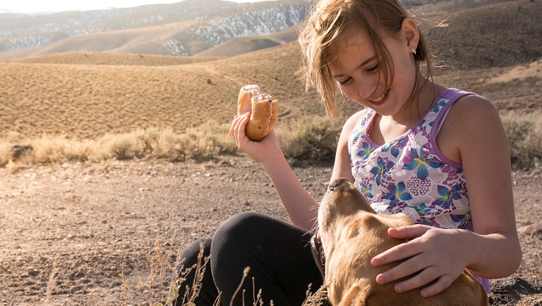 A girl smiles and pets her dog's head with one hand as she holds a bagel in the other hand.