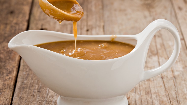 A close up shot of a small white ladle pouring brown gravy into a white ceramic gravy boat. Shot on a grungy old wooden table.