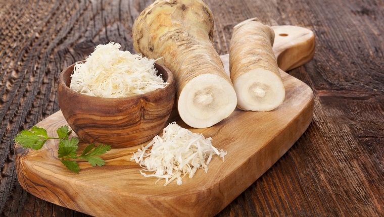 Fresh and grated horseradish in wooden bowl with parsley on wooden cutting board. Horse-radish root.