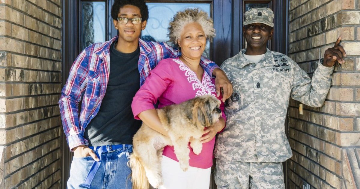 Black family of 3 members-including a son, mom, and father in a Armed Forces uniform-standing in doorway with mom holding their small dog.