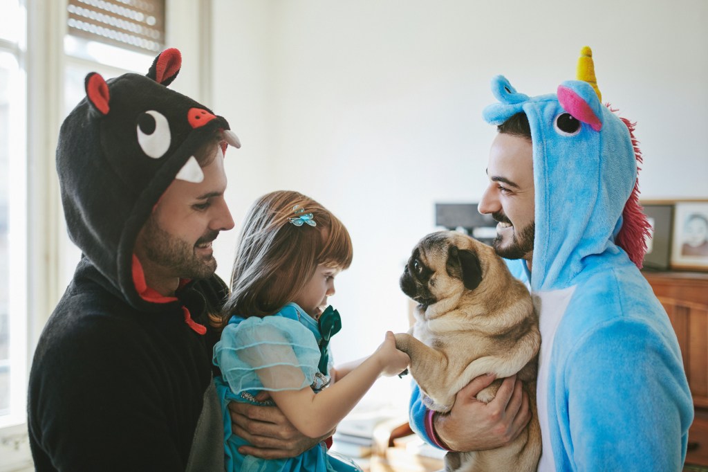 Happy gay couple in pj costumes carrying daughter and Pug. Family with pet is spending leisure time at home with their new dog they chose for adoption.