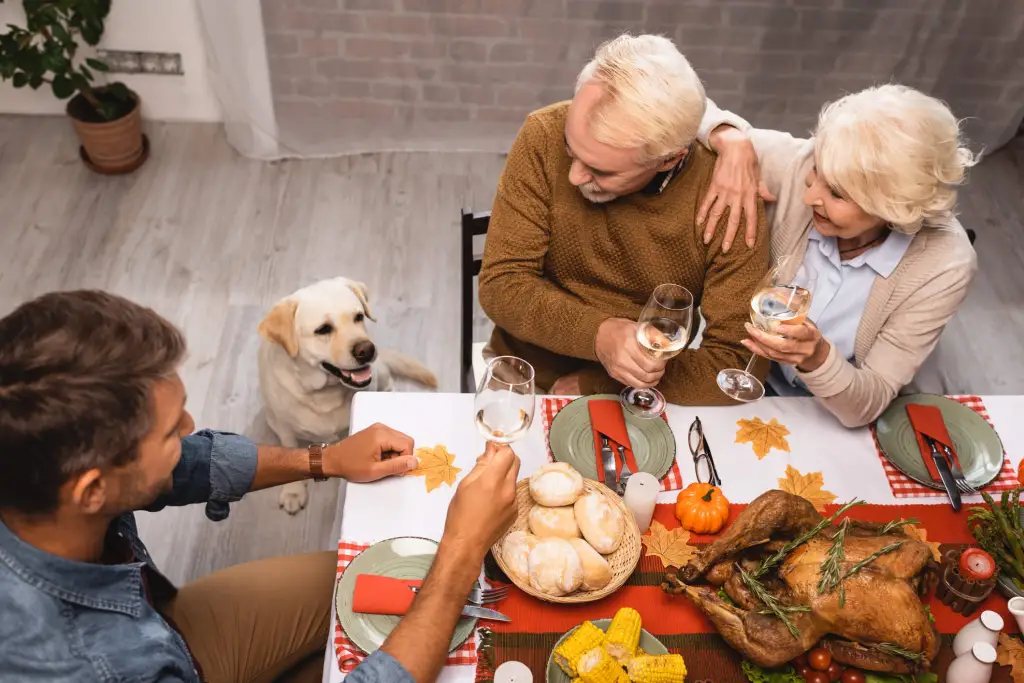 Family eating Thanksgiving dinner with rolls and turkey on table. Senior couple to the right of a yellow Lab. Man to the left of dog.