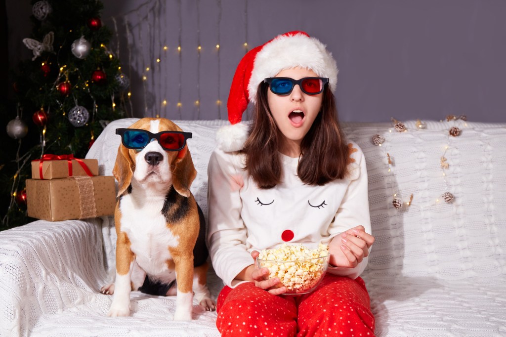 A girl in a Santa Claus hat and 3D glasses is sitting on the couch, watching Christmas movies with her Beagle.
