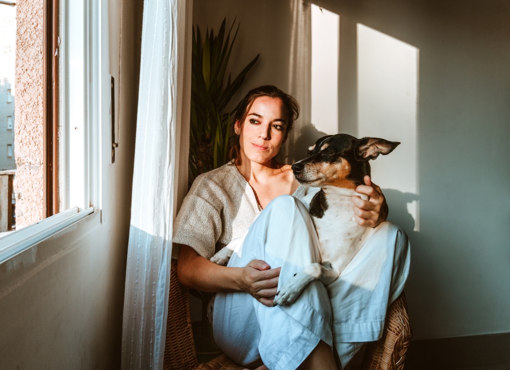 medium shot of a young woman with a white sweater sitting in a wicker chair in her apartment with her faithful dog