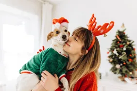 Smiling woman wearing reindeer antler headband holds her pup, who she dressed in a Santa hat and festive sweater as a holiday tradition to share with her dog.