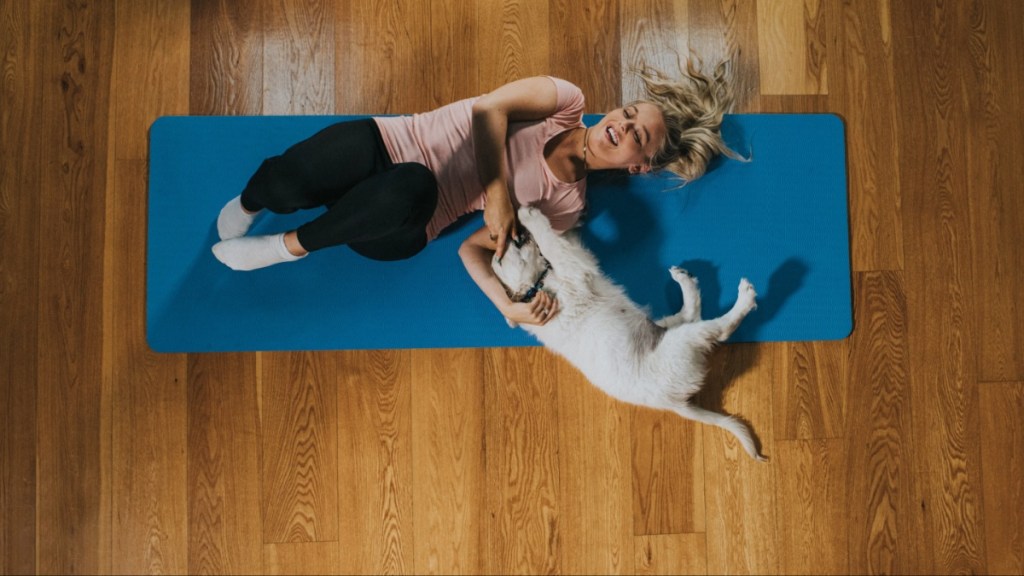 Young woman and a playful golden retriever puppy lie on a yoga mat on a wooden floor, next to each other. She lies on her back and he lies on his side looking at her. She looks back at him and smiles.