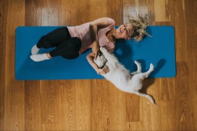 Young woman and a playful golden retriever puppy lie on a yoga mat on a wooden floor, next to each other. She lies on her back and he lies on his side looking at her. She looks back at him and smiles.