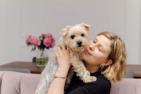 Young woman embraces a Yorkshire Terrier — a small, cuddly dog breed — while sitting on pink sofa in cozy modern living room.